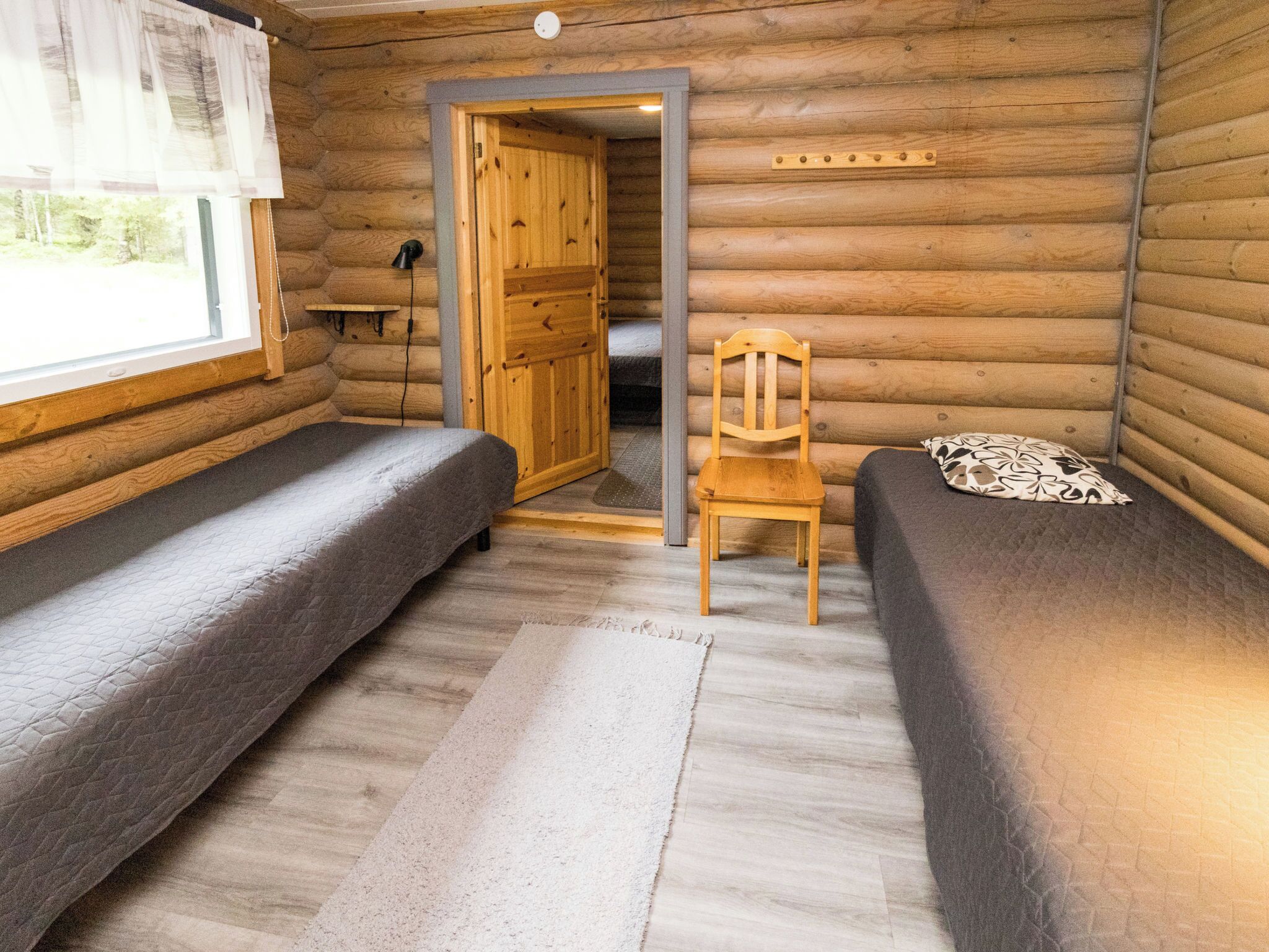 Habitació d'una cabana de fusta amb dos llits, una cadira de fusta i una porta que fa a l'exterior.