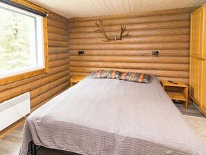 A log cabin bedroom with a bed, pillows, a nightstand, a heater, and a window.
