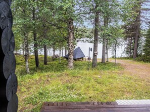 Uma cabana com telhado de palha, rodeada por árvores, uma área gramada e um corpo de água ao fundo.