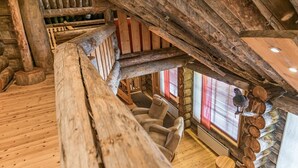 Ein rustikales Blockhaus-Interieur mit einer Holztreppe, einer gemütlichen Sitzecke und einem großen Fenster mit roten Jalousien.