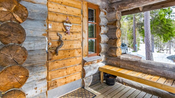 Eine Blockhütte mit Holztür, einer Bank und einem Fenster mit Blick auf eine verschneite Landschaft.