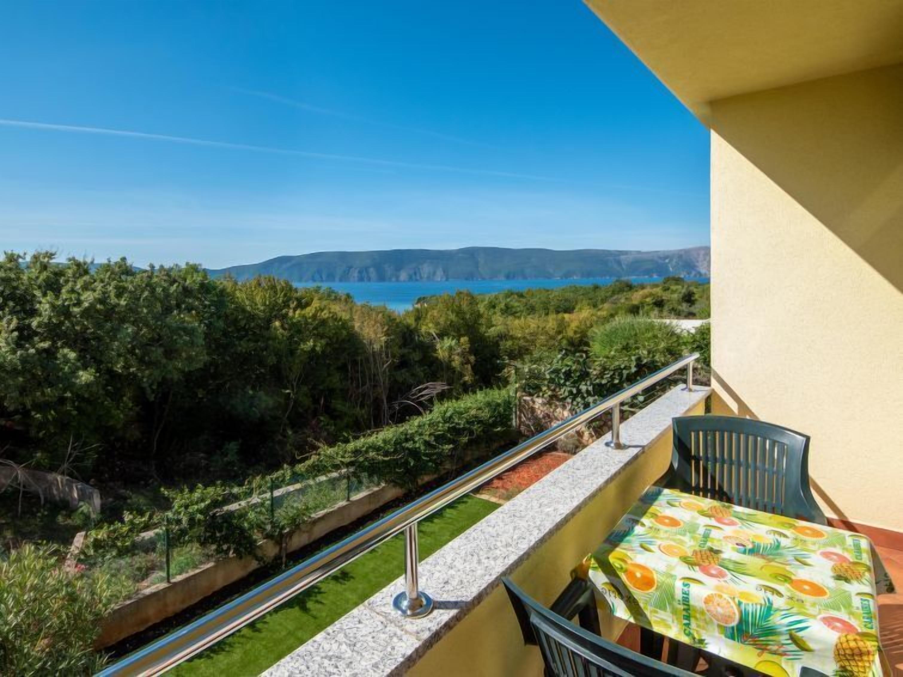 A balcony with a table and chairs, overlooking a coastal landscape with greenery and a view of the sea and distant hills.