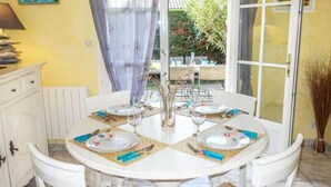 Mesa de jantar posta para quatro pessoas, com cadeiras brancas, um arranjo de flores no centro e vista do jardim através de uma porta de vidro.