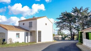 Uma casa branca de dois andares com telhado de telhas, garagem e entrada com portão, acessível por um caminho.
