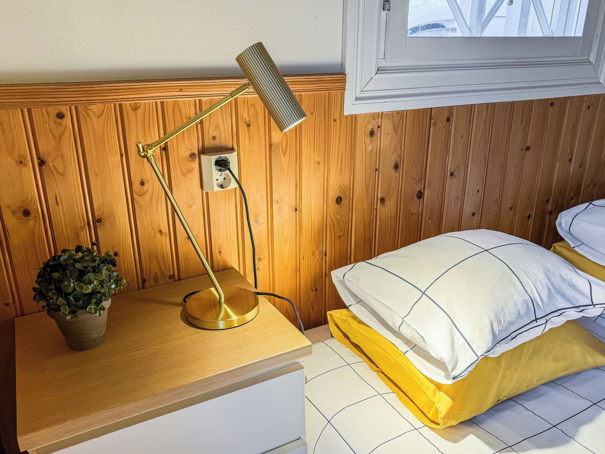 A wooden-paneled room with a bedside table, a lamp, a potted plant, and a bed with a white and yellow bedding.
