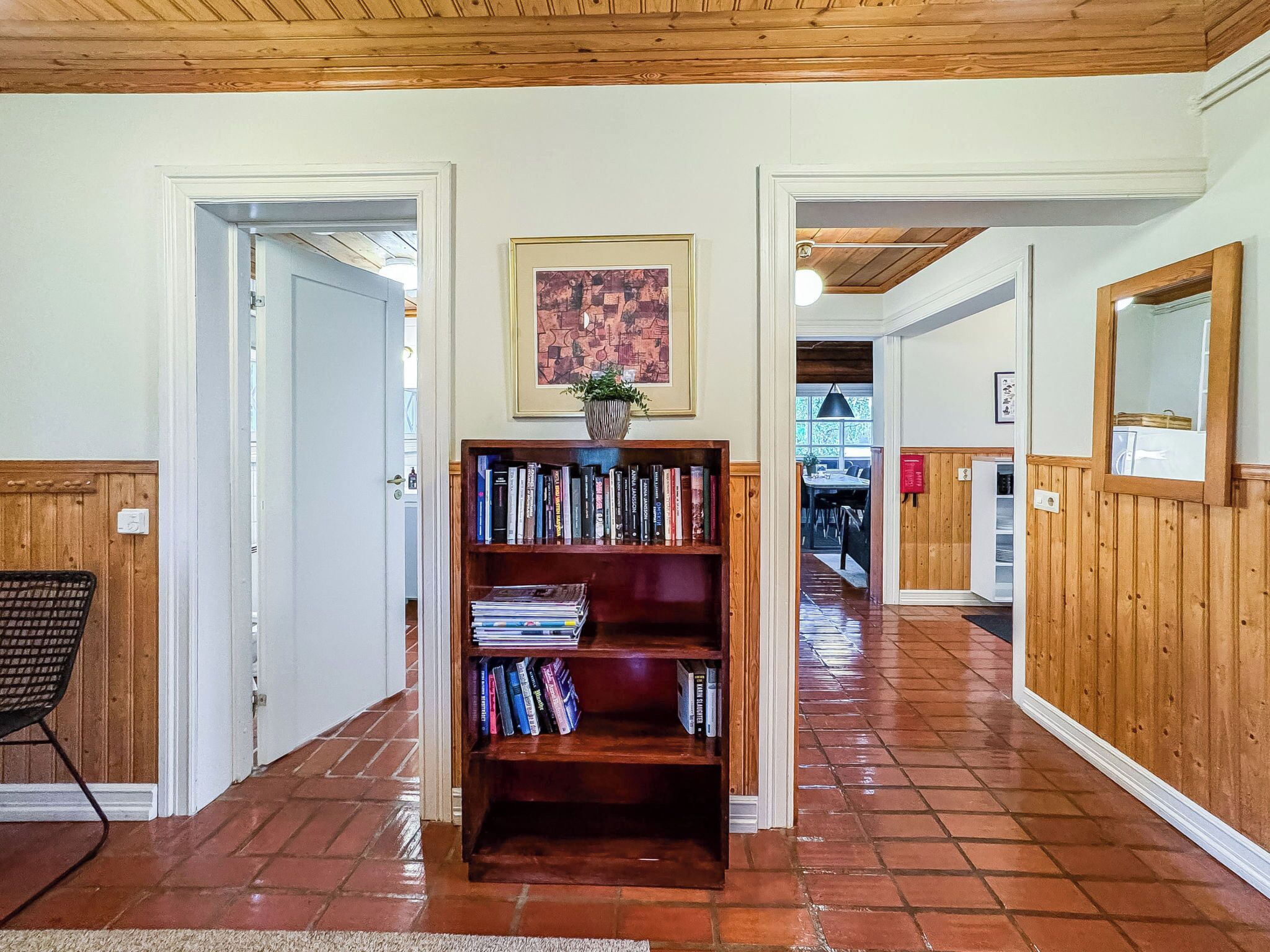 A hallway with a wooden bookshelf, a framed picture, and a dining area in the background.