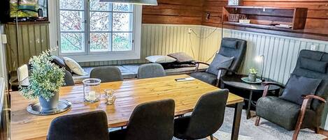 A wooden cabin interior with a dining table, chairs, a hanging lamp, a window with a view of trees, and a shelf with items.