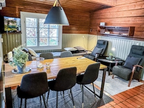 Ein Interieur einer Holzhütte mit Esstisch, Stühlen, einer Hängelampe, einem Fenster mit Blick auf Bäume und einem Regal mit Gegenständen.