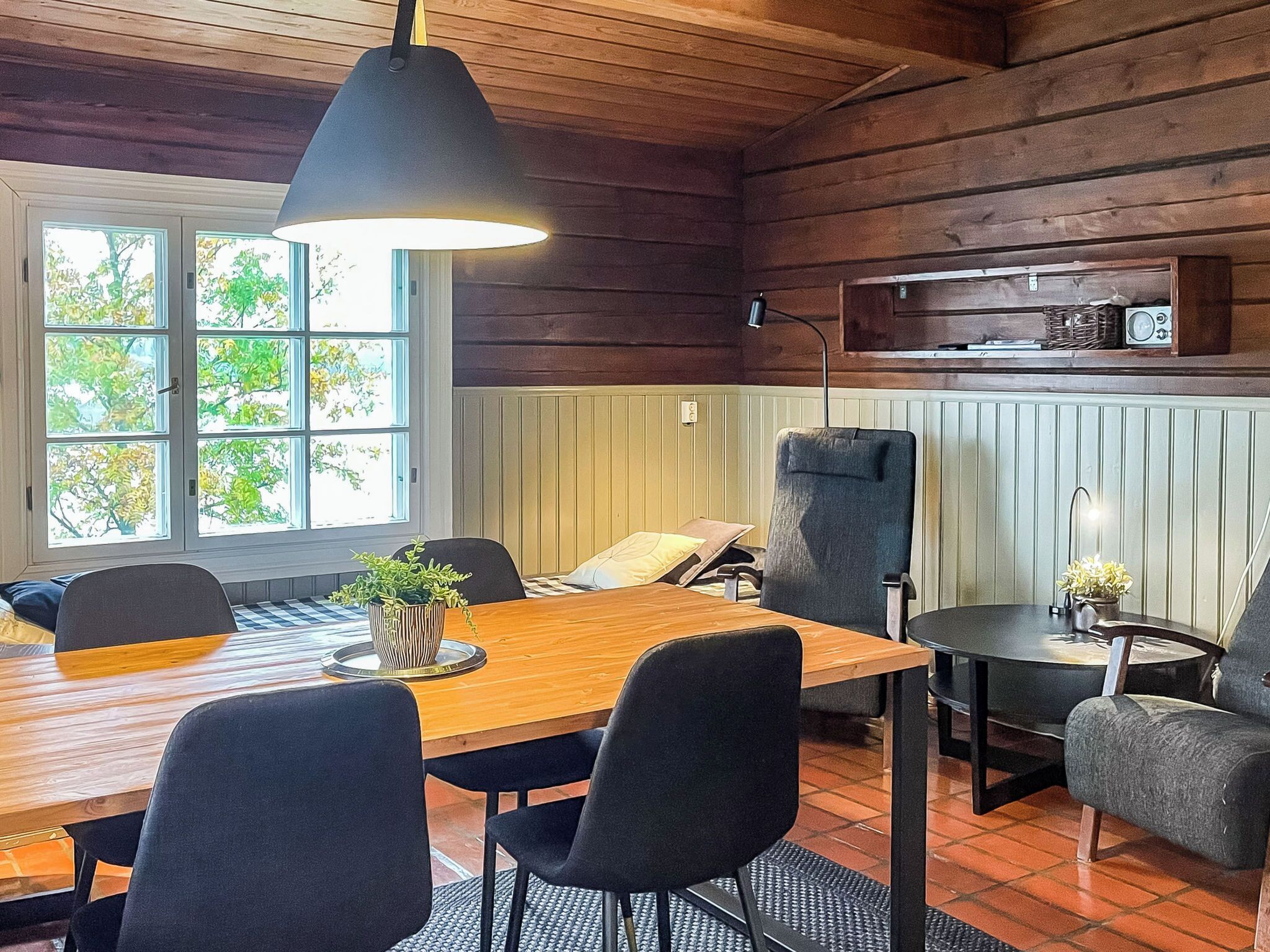 Un intérieur en bois comprenant une table à manger, des chaises, un luminaire suspendu, une petite table d’appoint et une fenêtre donnant sur des arbres.
