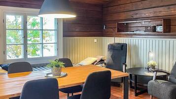 Un intérieur en bois comprenant une table à manger, des chaises, un luminaire suspendu, une petite table d’appoint et une fenêtre donnant sur des arbres.