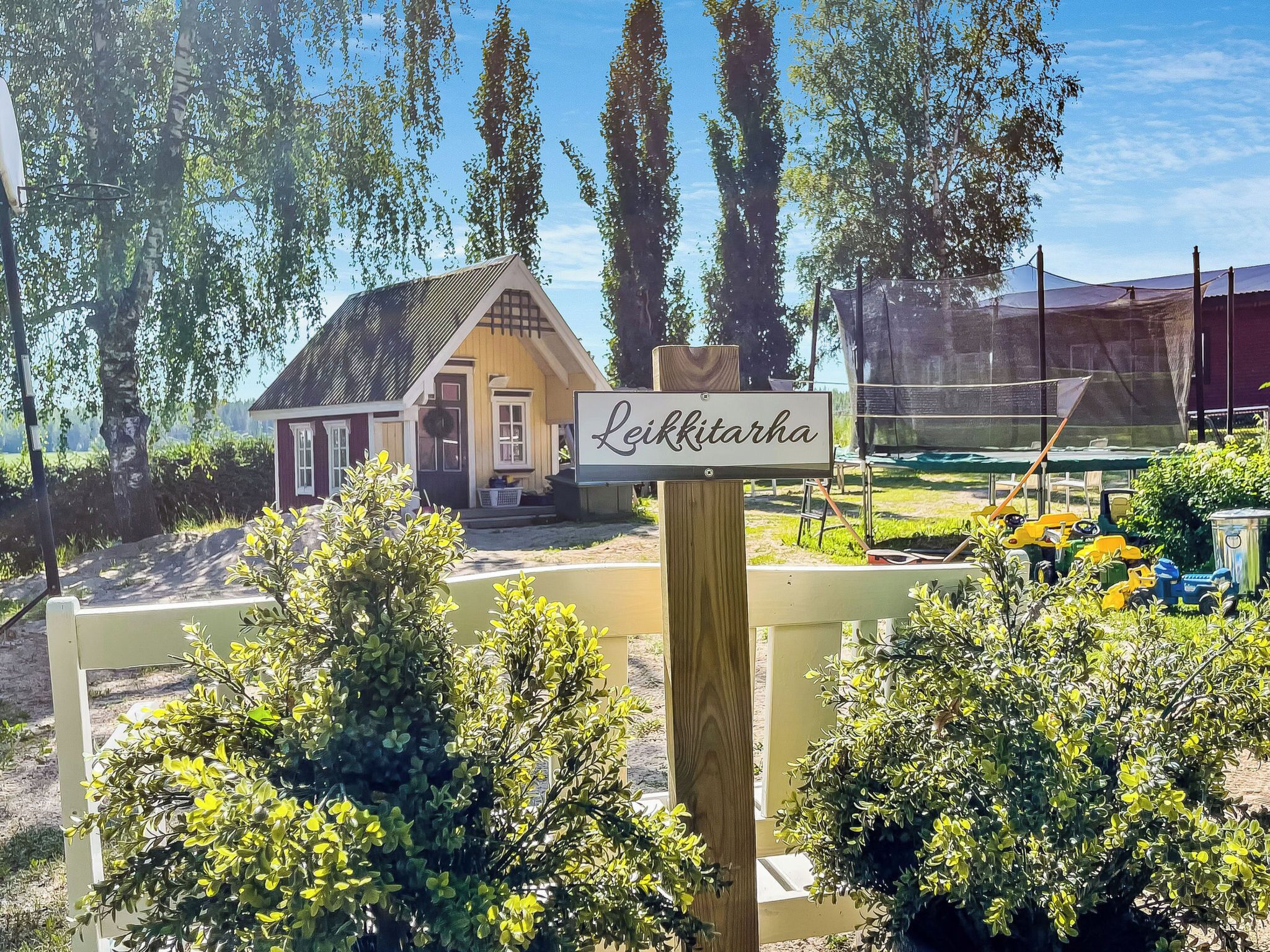 Una casa pequeña con un cartel que dice "Leikkitarha" en un jardín con árboles y una valla.