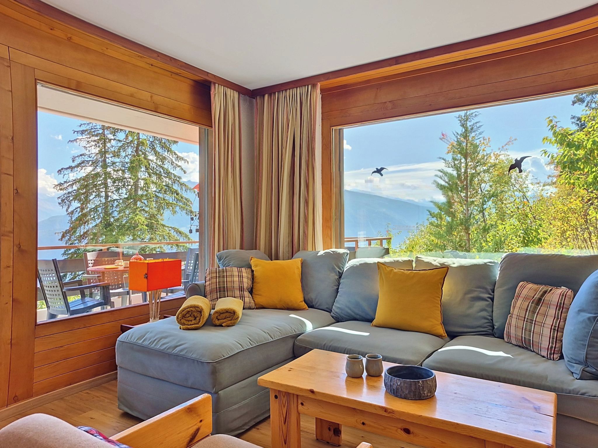 Una sala de estar moderna con un sofá modular, suelo de madera y ventanas amplias con vistas a árboles y montañas.