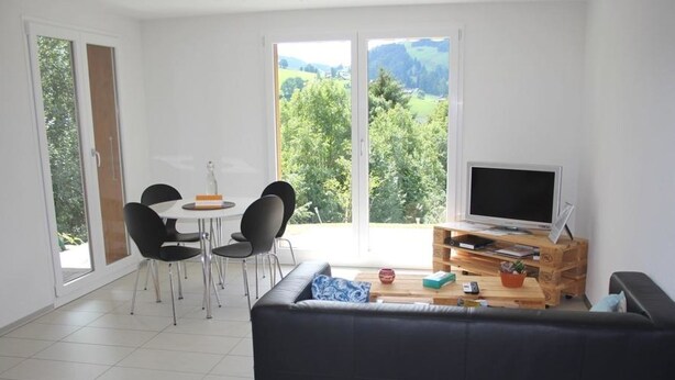A modern living room with a black sofa, a dining table with chairs, a TV on a wooden stand, and large windows offering a view of greenery.