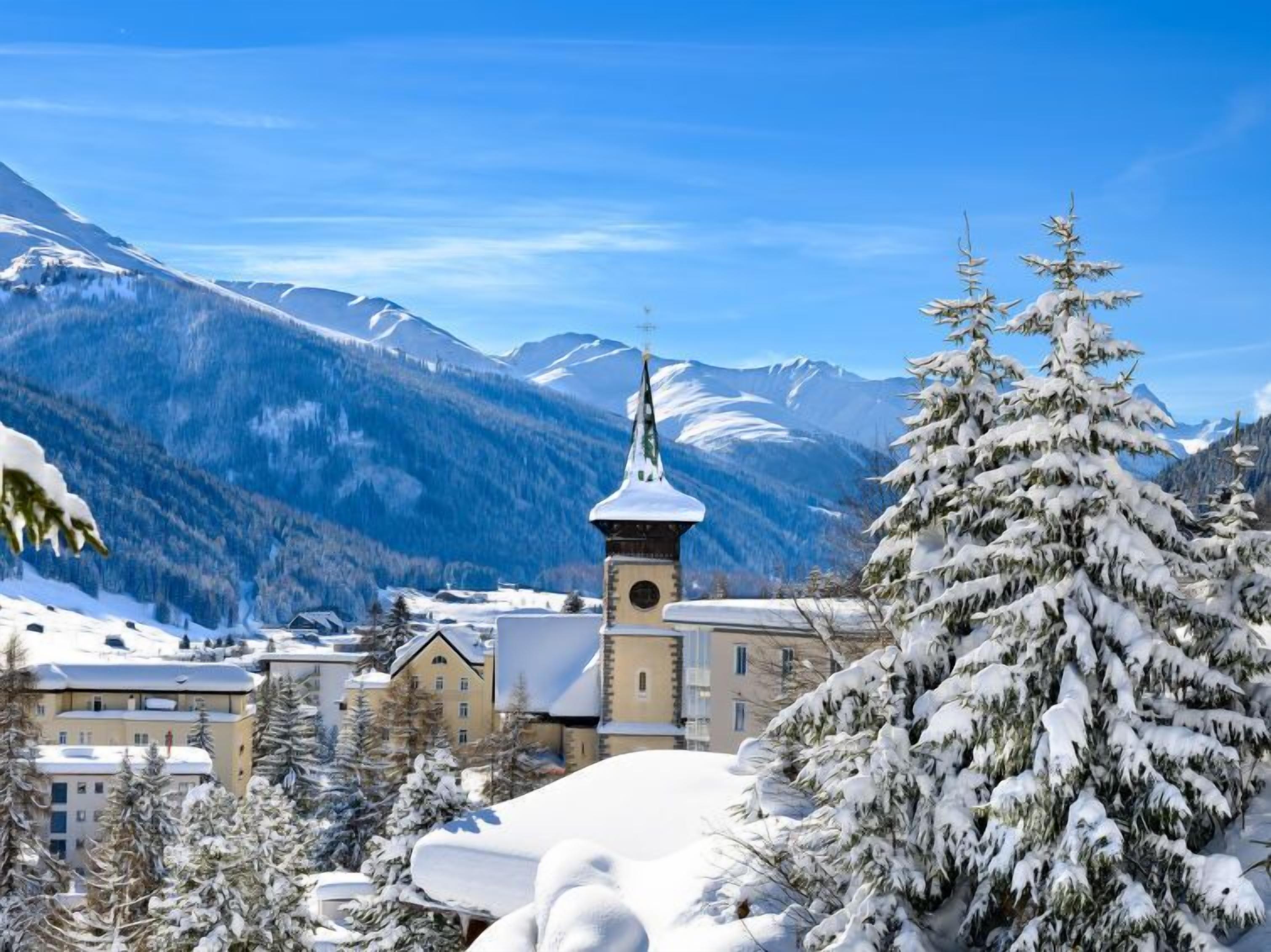 Eine verschneite Bergstadt mit einem Kirchturm, Wohnhäusern und Schnee bedeckten Bäumen.