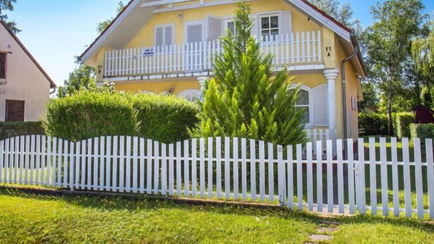 A two-story yellow house with a white picket fence, a balcony, and a well-maintained lawn. - Vacation home Welle by Interhome (Balatonmariafurdo)