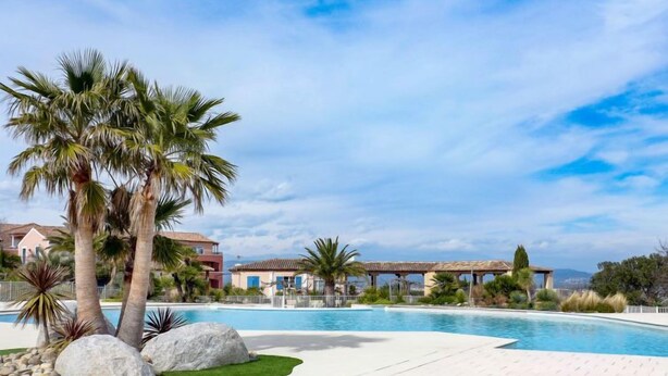 A swimming pool with palm trees and a thatched-roof structure in the background. - Le Golf de Roquebrune by Interhome (Roquebrune sur Argens)