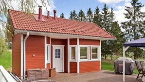 Punainen puinen rakennus, jossa on tiilikatto, ovi ja ikkunat. Puisen terassin yhteydessä on grilli ja ulkotila, jossa voi istua.