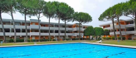 A swimming pool surrounded by multi-story buildings with balconies, trees, and a clear sky.