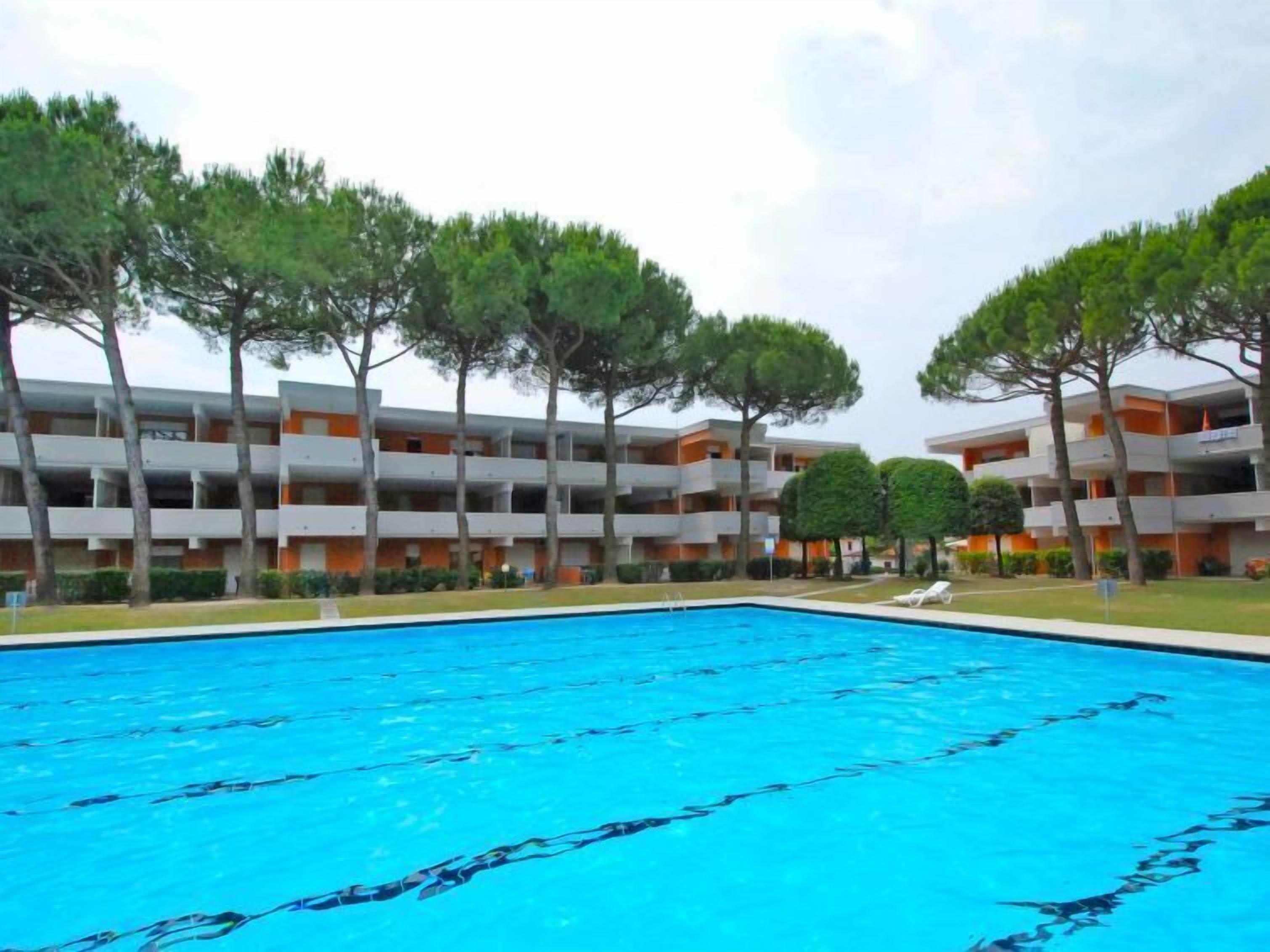 A swimming pool surrounded by multi-story buildings with balconies, trees, and a clear sky.