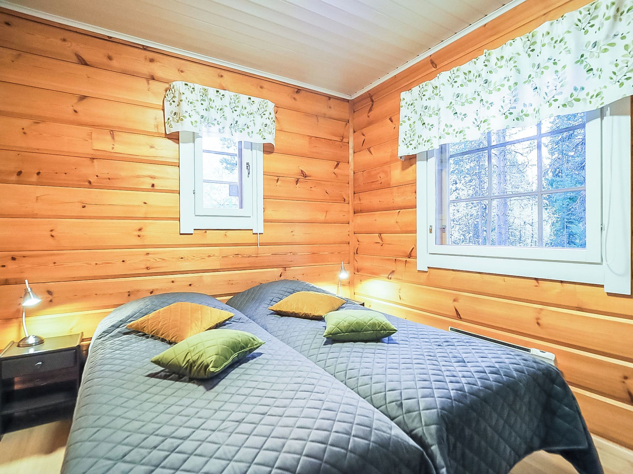 Une chambre dans un chalet en bois, avec deux lits, une table de chevet, une lampe et une fenêtre donnant sur la forêt.