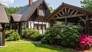 Une maison traditionnelle à colombages, dotée d’un porche en bois et d’un jardin agrémenté de plantes à fleurs.