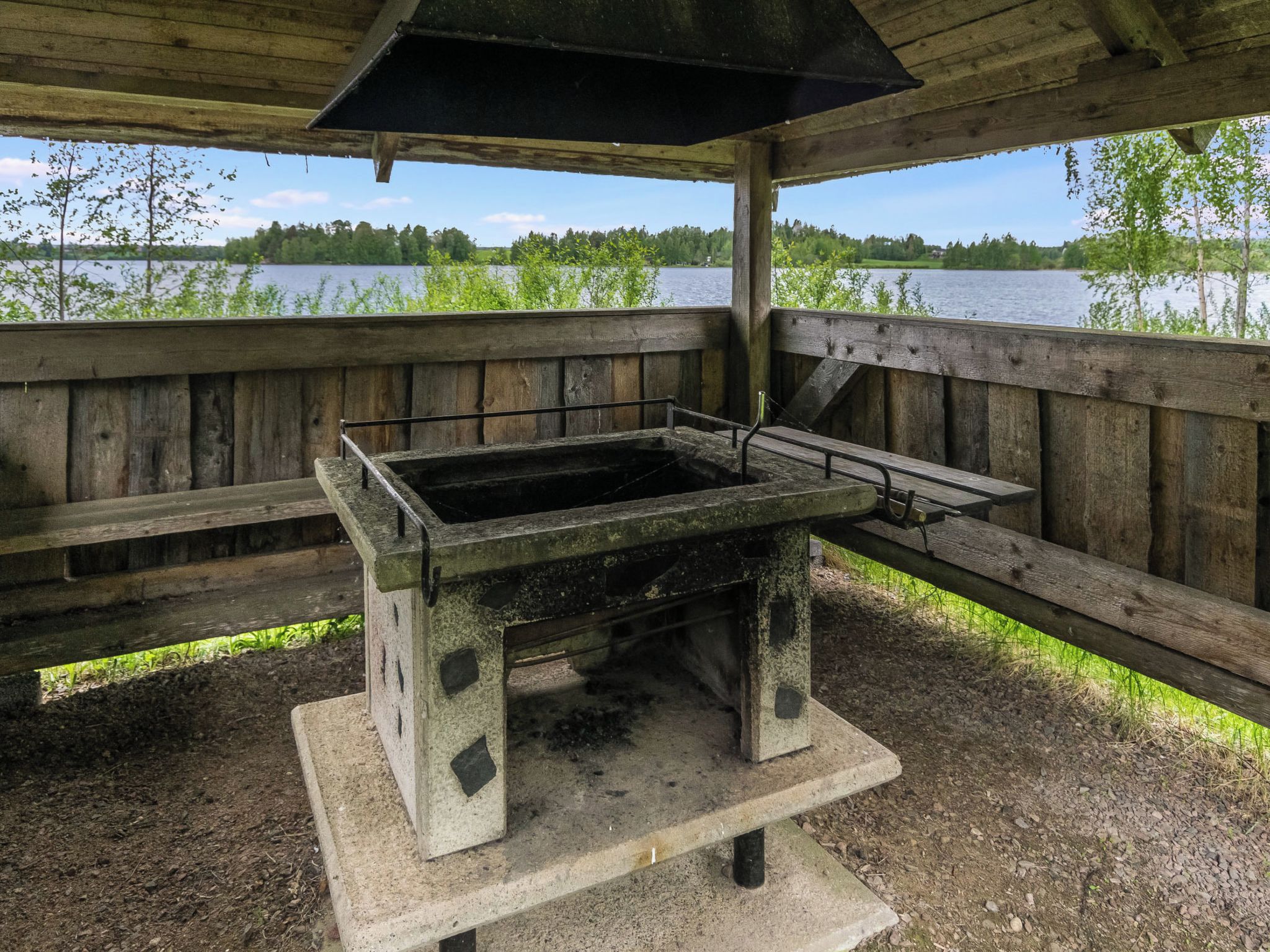 A rustic outdoor grill area with a concrete table and benches, surrounded by wooden railings, overlooking a lake.