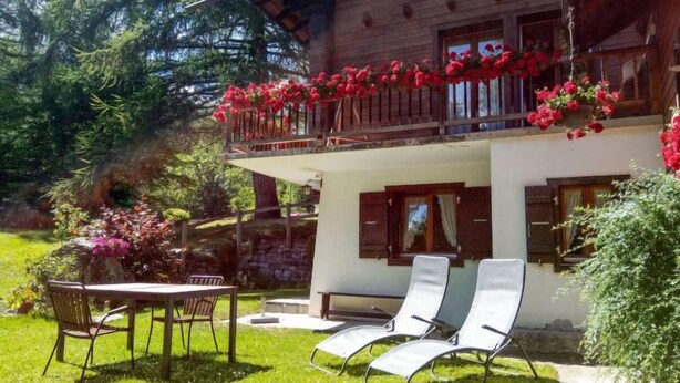 A wooden house with a balcony adorned with red flowers, a dining area with a table and chairs, and a garden with green grass and shrubs. - Le Tzanton by Interhome (Ovronnaz)