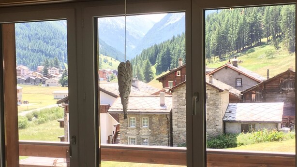 Depuis la fenêtre, on aperçoit un village de montagne avec ses bâtiments traditionnels en pierre, une clôture en bois et une mangeoire à oiseaux suspendue.