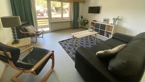 Un salon moderne avec un canapé en cuir noir, une chaise en bois, une petite table et un téléviseur fixé au mur.