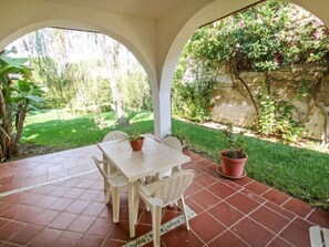 A covered patio with a table and chairs, surrounded by a garden with a stone wall and potted plants.