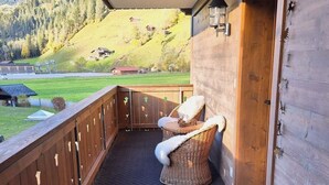 Ein hölzerner Balkon mit einem Korbstuhl und einem kleinen Tisch, der auf eine grüne Landschaft mit Hügeln und ein Gebäude mit rotem Dach im Hintergrund blickt.
