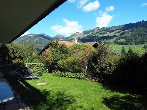 Blick von einer überdachten Außenbereits mit Tisch und Stühlen auf eine gebirgige Landschaft mit einem kleinen Gebäude und üppiger Vegetation.