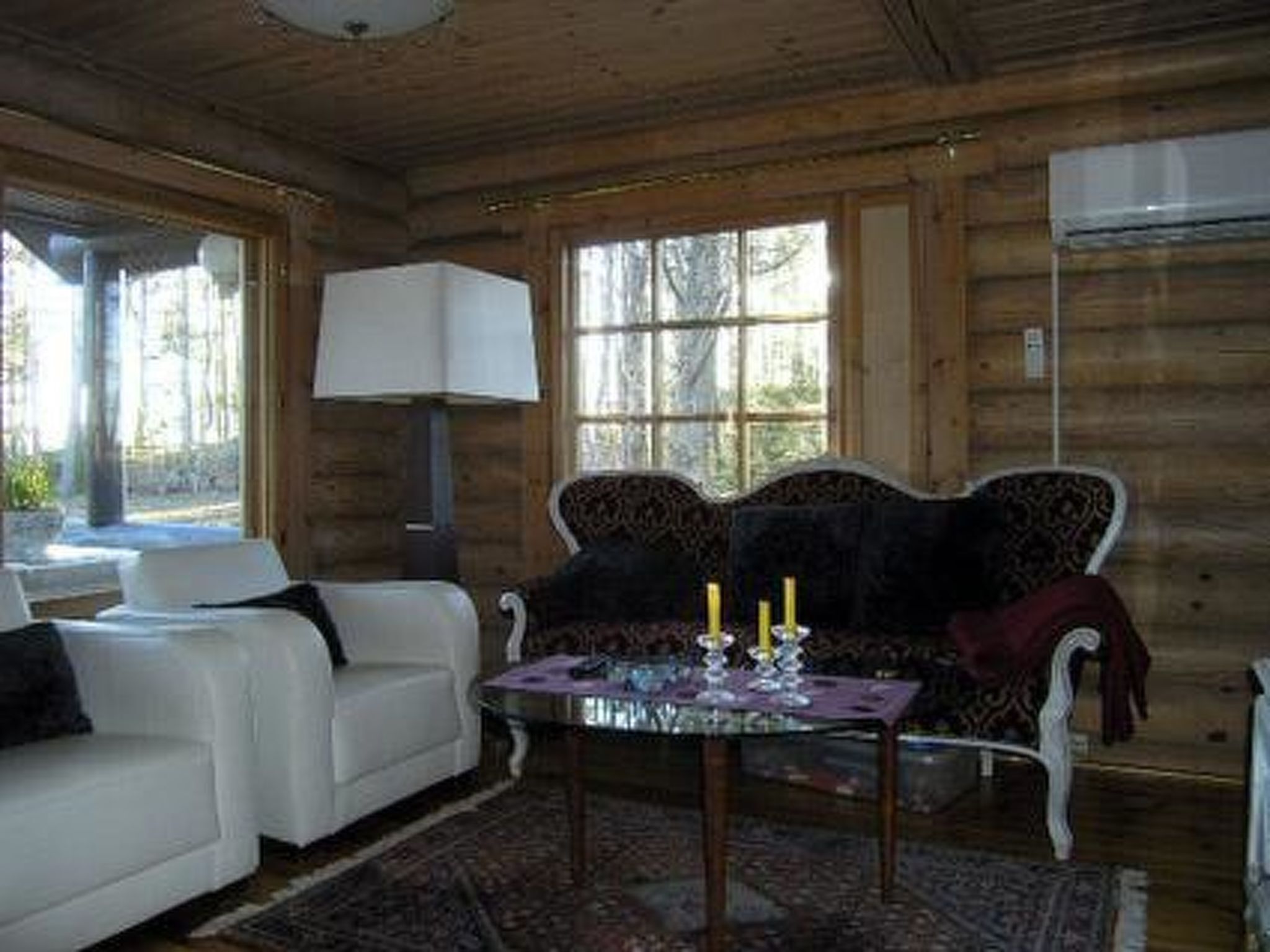 Het interieur van een houten hutje met een witte bank, een gestreept fauteuil, een glazen salontafel en een raam met uitzicht op bomen.