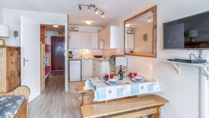 Una cocina compacta con armarios blancos, microondas y un refrigerador pequeño. Hay una mesa de comedor con vasos rojos y un banco. Un televisor está montado en la pared.