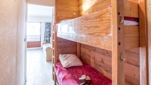 Una cama litera con cabecera de madera, una cama individual con una manta rosa y un almohadón, y un par de zapatillas en el suelo.