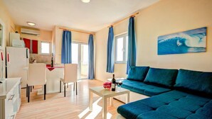 A modern living room with a blue sofa, a dining area with a white table and chairs, and a kitchen area with a refrigerator and an air conditioner. - Melandura by Interhome (Pag/Dinjiška)