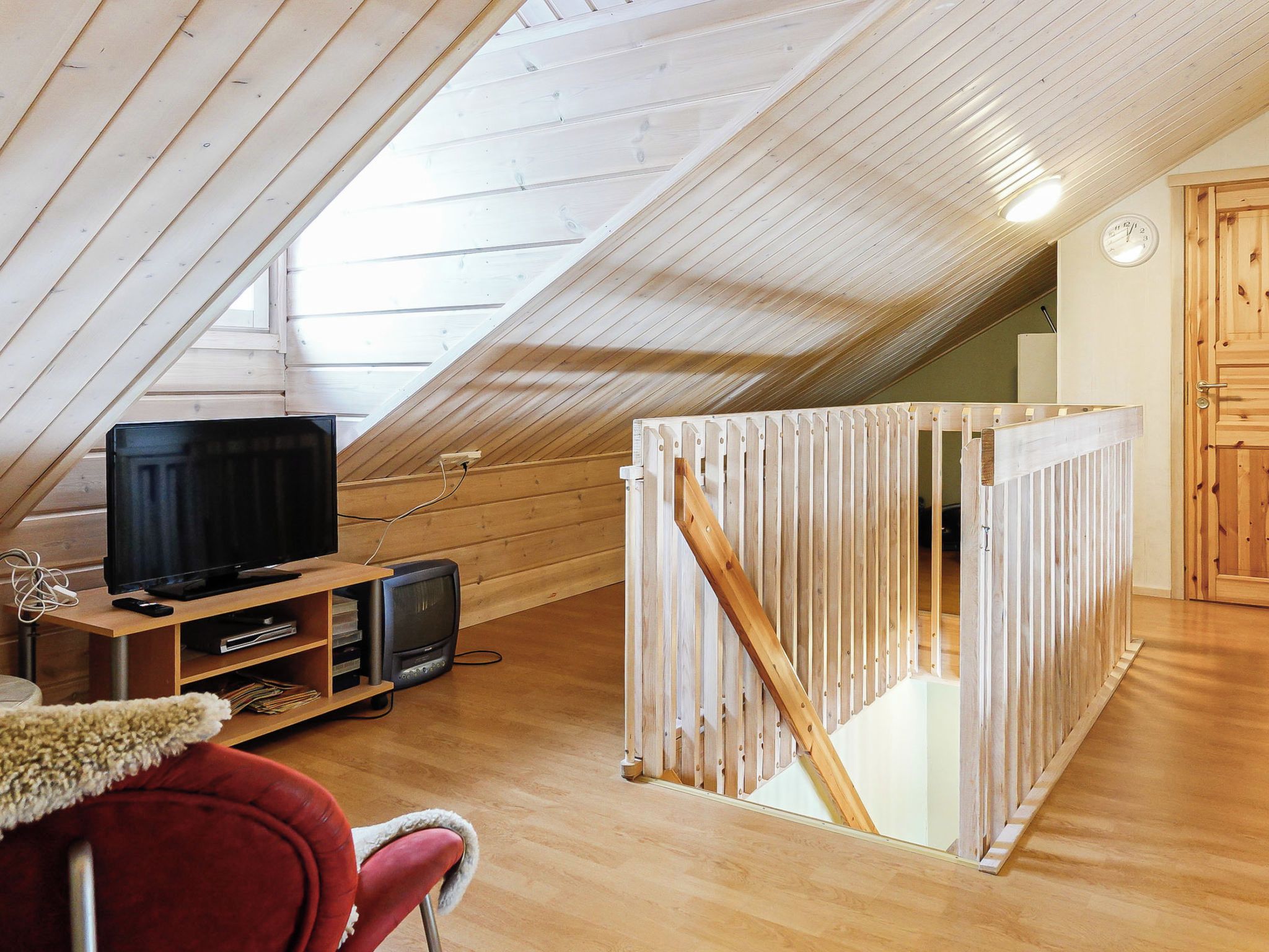 A room with a sloped ceiling, wooden flooring, a television, a small television, and a red chair.