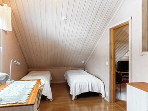 A small, single-room accommodation with two beds, a desk with a lamp, and a wooden ceiling.