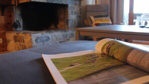 A room with a stone fireplace, a table, and a magazine on a cushion. - Ambassadeur 15 by Interhome (Villars)
