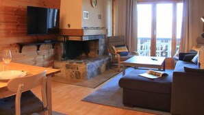 A cozy living room with a stone fireplace, a flat-screen TV mounted on the wall, a comfortable sofa, a dining table with chairs, and a view of the outside through large windows. - Ambassadeur 15 by Interhome (Villars)