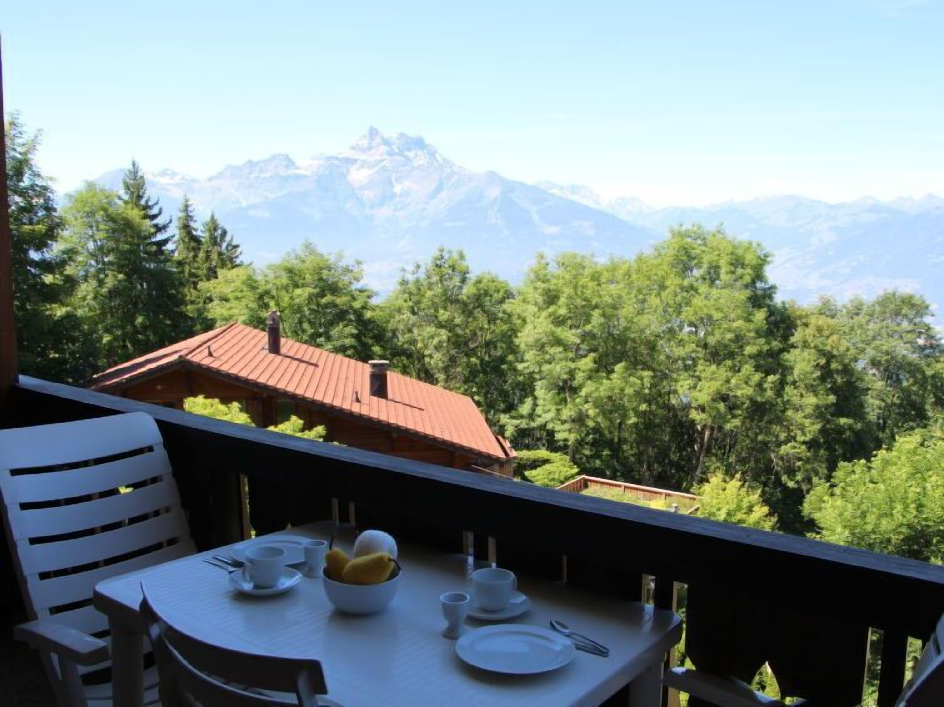 A dining table set with plates, bowls, and utensils, overlooking a mountainous landscape with trees and a red-roofed building.