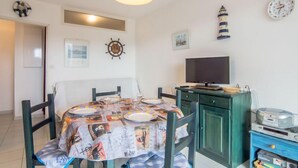 Un coin repas avec une table ronde recouverte d’une nappe à motifs, entourée de chaises. Un téléviseur est fixé au mur, et divers éléments décoratifs ornent les murs, notamment un gouvernail de navire et des tableaux encadrés.