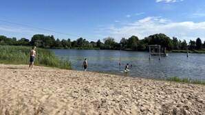 Una spiaggia sabbiosa con un lago, un piccolo molo e alberi sullo sfondo.