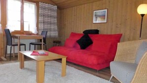 A cozy room with a red sofa, a wicker chair, a wooden table, and a dining area with chairs and a table. - Chalet Doris by Interhome (Grindelwald)