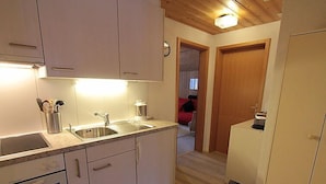 A compact kitchen with white cabinets, a sink, a stove, and a microwave. There is a wooden ceiling and a visible clock on the wall. - Chalet Doris by Interhome (Grindelwald)