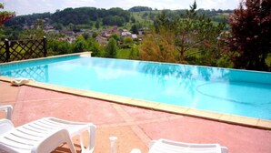 Ein Swimmingpool mit klarem, blauem Wasser, umgeben von einer gefliesten Terrasse. Weiße Kunststoffliegen und ein kleiner Tisch sind vorhanden. Im Hintergrund bietet sich ein Blick auf eine grüne Landschaft mit Bäumen und Häusern.