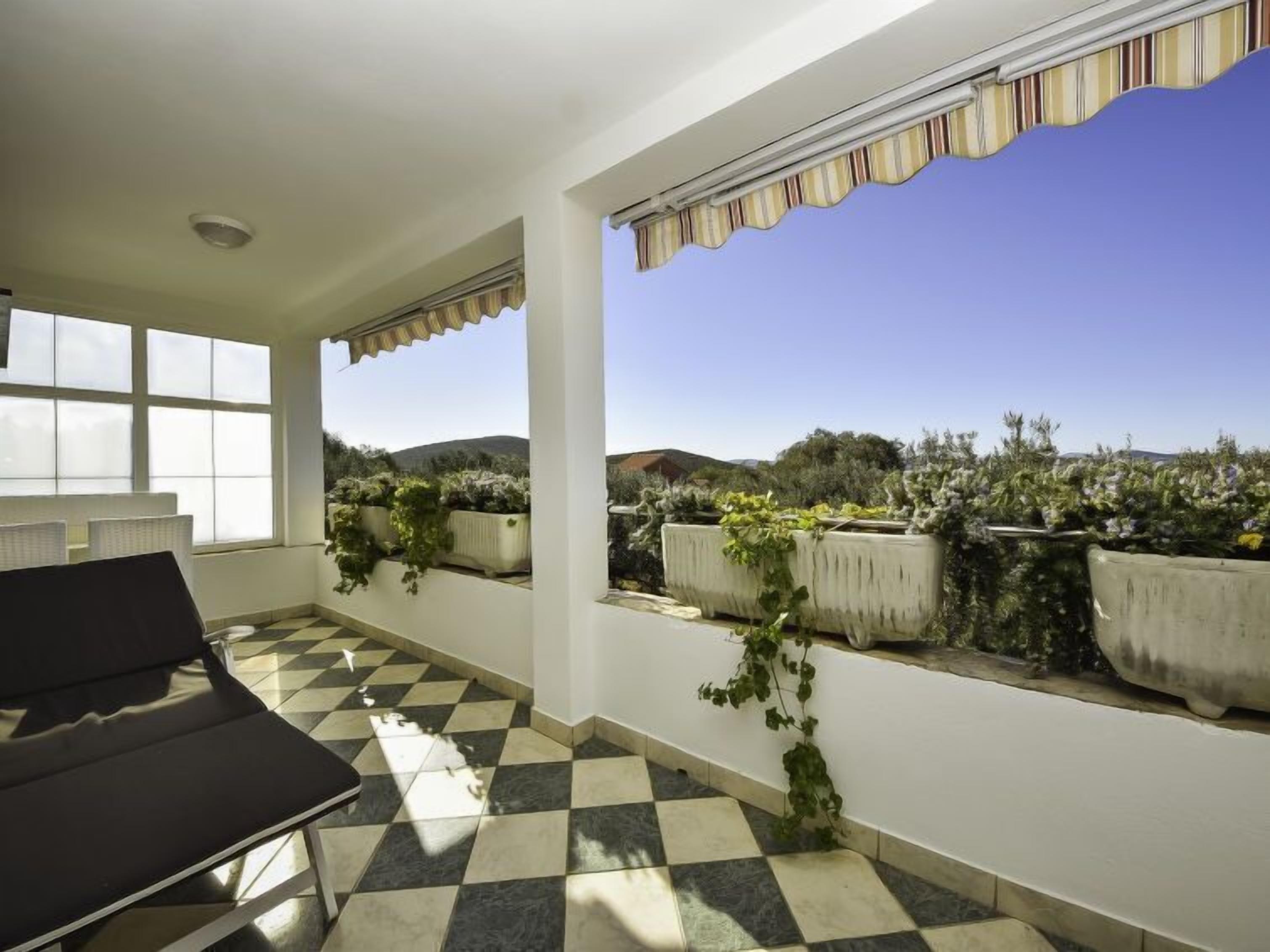 Una habitación iluminada por el sol, con piso a cuadros, una silla negra y vistas a plantas en macetas y colinas al exterior.