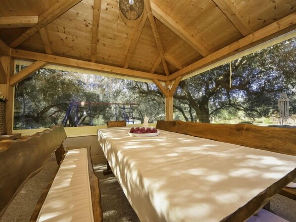 Un coin repas en bois avec une longue table et des bancs, de grandes fenêtres donnant sur la forêt et un luminaire suspendu.