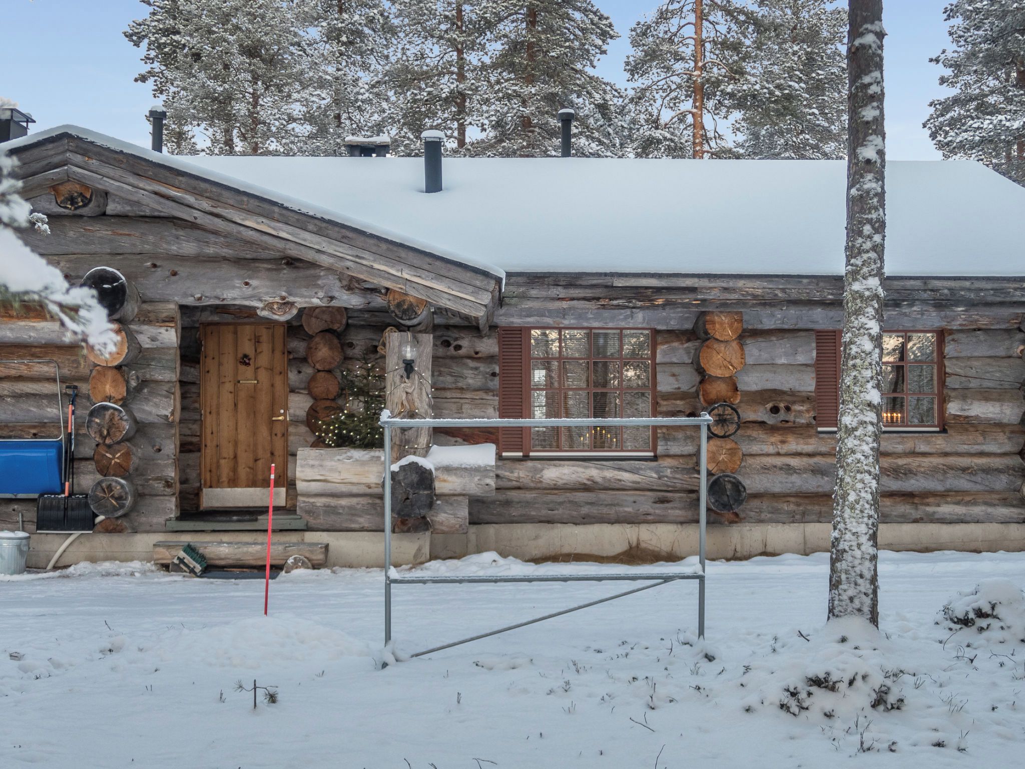 Lumipeitteinen mökki, jossa on puinen ovi ja metallinen portti edessä.
