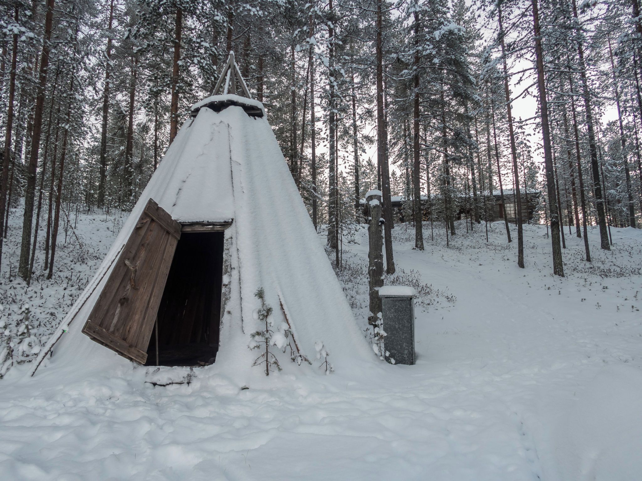A snow-covered wooden structure with an open door, surrounded by a forest.
